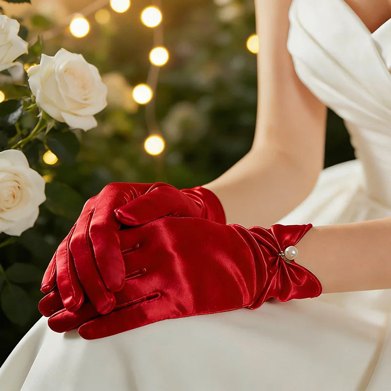Gants femme en satin rouge portés, mains croisées devant des roses blanches